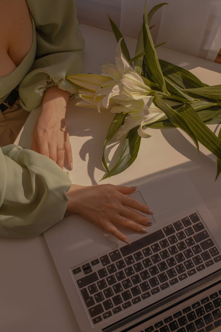 Woman's hands on a laptop with white lilies on a desk, bathed in warm indoor lighting.